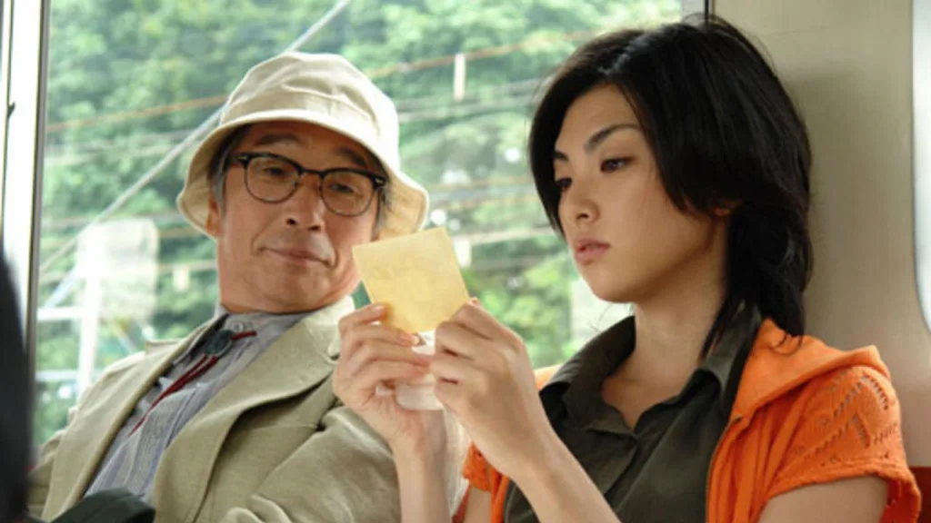 Cena do filme "Yunagi City, Sakura Country". Um homem e uma mulher sentados em um transporte público. O homem usa chapéu e óculos, enquanto a mulher lê uma nota autoadesiva, ambos com expressões concentradas.