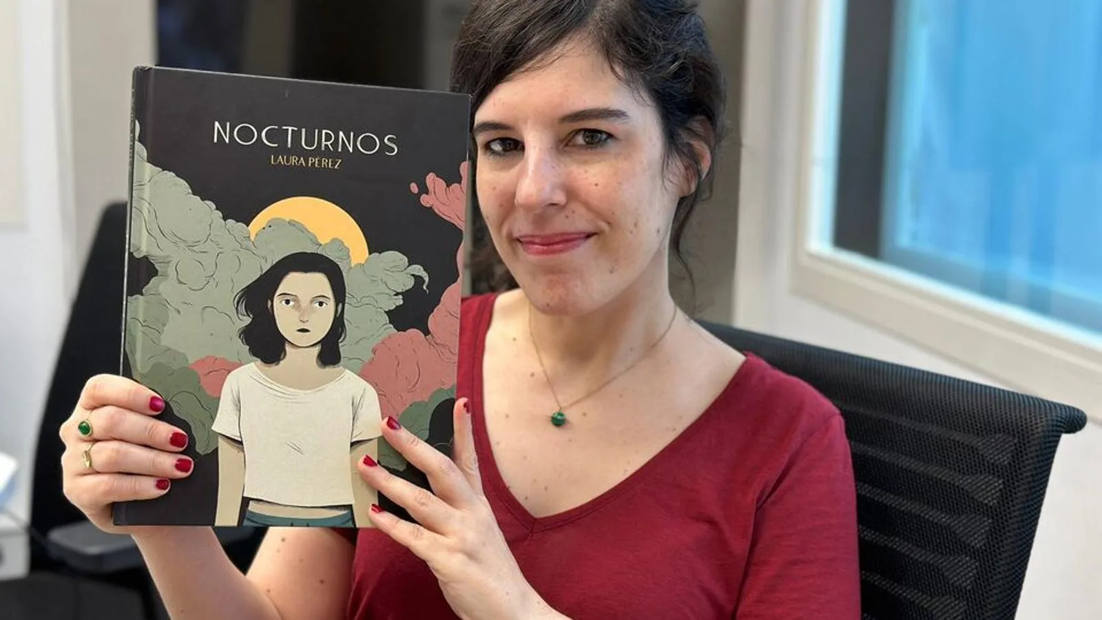 Laura Pérez, uma mulher com cabelo escuro e roupa vermelha, segura o quadrinho "Noturnos", sorrindo.