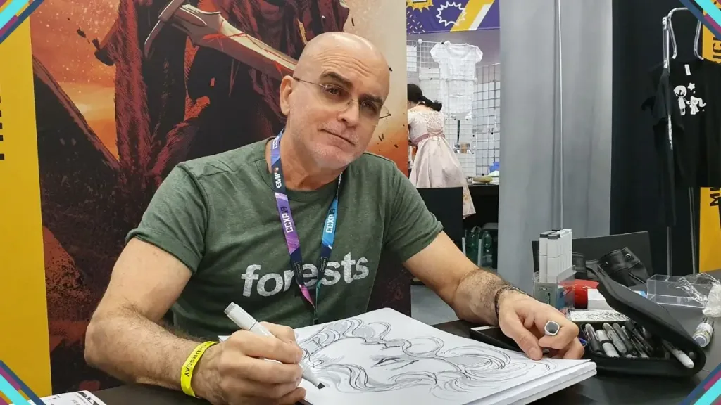 Homem sentado em uma mesa, assinando um desenho com caneta, em evento de quadrinhos ou cultura pop, com fundo colorido e outros stands ao redor.