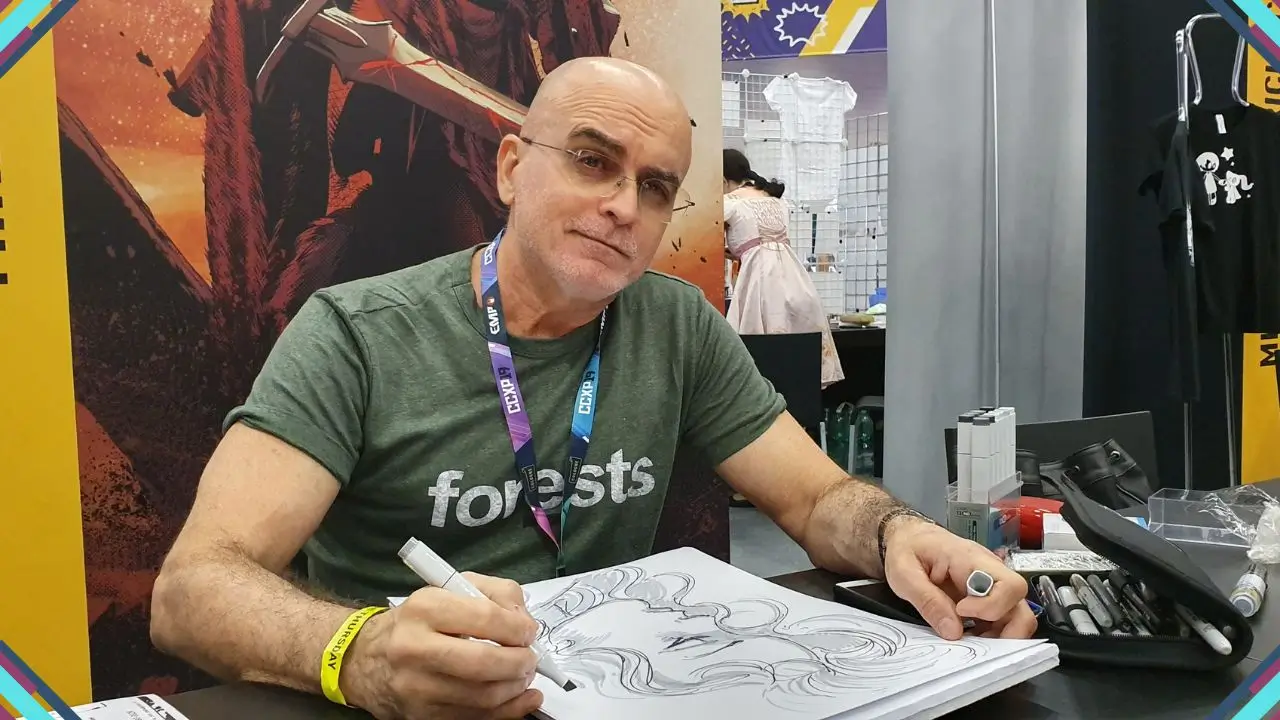 Homem sentado em uma mesa, assinando um desenho com caneta, em evento de quadrinhos ou cultura pop, com fundo colorido e outros stands ao redor.